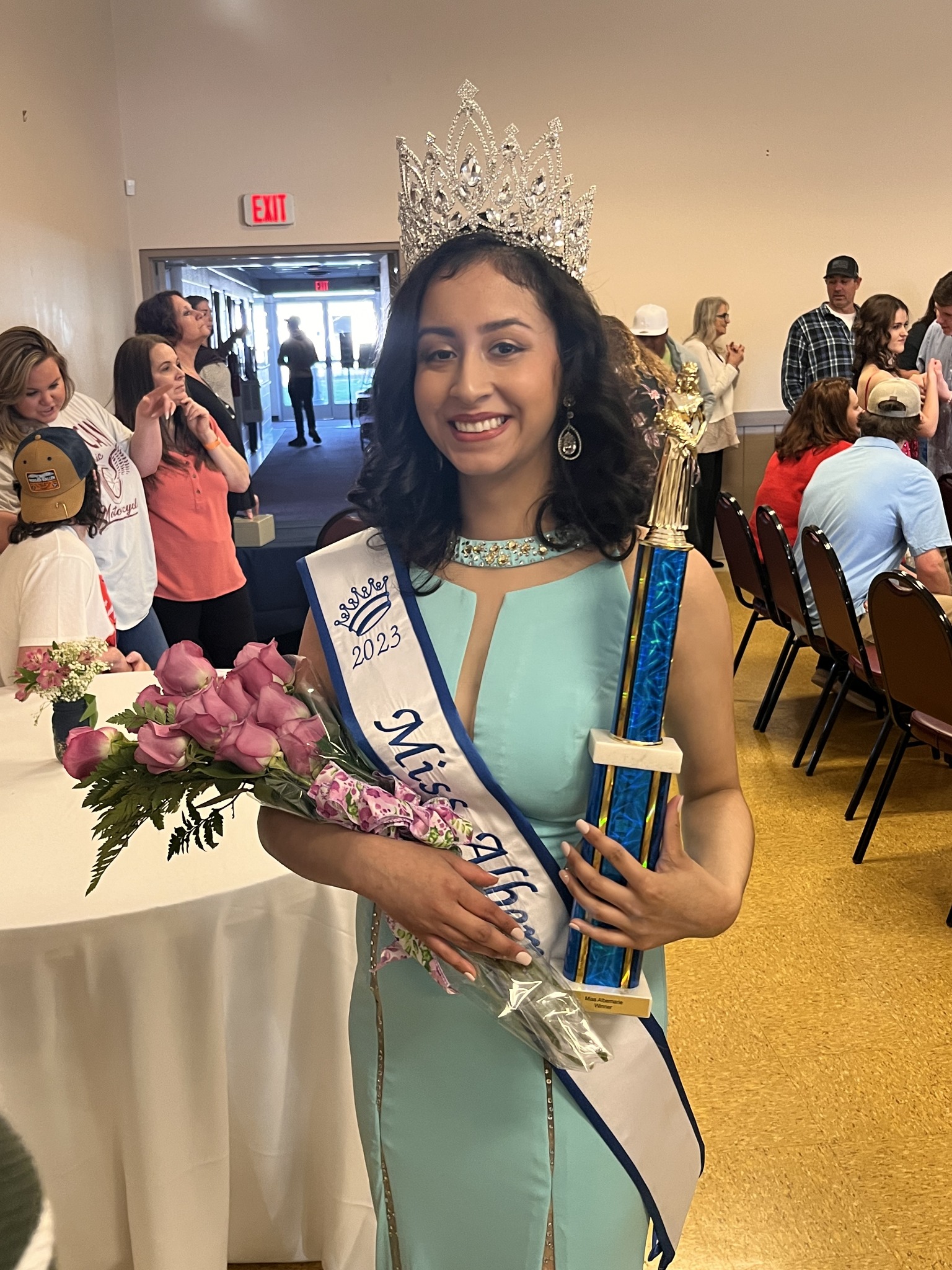 Crowned contestant in an aqua evening gown holding flowers and a trophy