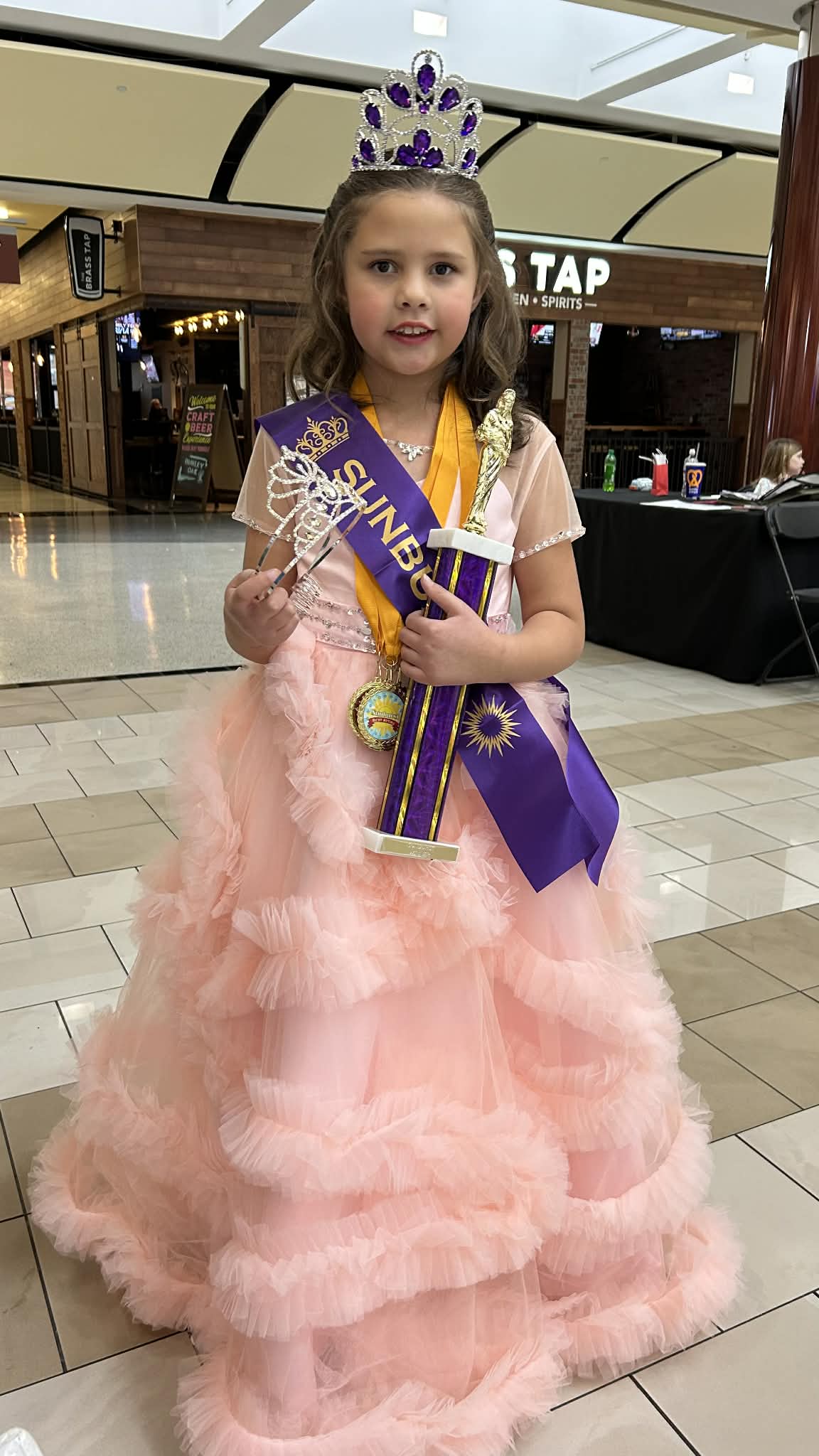 Crowned contestant in a peach evening gown holding a trophy